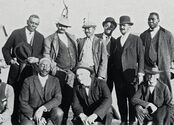 Members of the Colored Citizens Relief Committee and East End Welfare Board pose for a photo following the 1921 Tulsa Race Massacre. The individuals standing include the following (left to right): Williams, Phillips, Esta "Essie" A. Loupe, Reverend E. N. Bryant, Horace T. Hughes, and McLean. The men kneeling include the following (left to right): Perry R. Russell, Ottaway W. Gurley, Bush, and Tucker Gilmer.