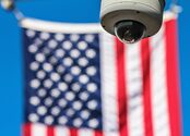A surveillance camera in the foreground, in front of a blurred out American flag in the background.