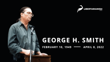 George H. Smith stands behind a podium against a black background. Text reads "George H. Smith - February 10, 1949 - April 8, 2022."