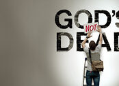 A man places a sign that reads 'NOT' in red, capital letters, on top of a wall that has the message 'God's Dead' painted on it to change the meaning of the sentence.