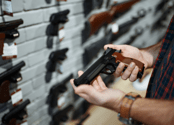 Various guns are displayed on a wall behind a man's hands holding a pistol. 