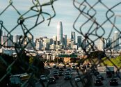 A highway and city skyline are seen through a hole in a chain link fence.