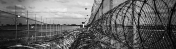 Black and white photograph with barbed wire on a chainlink fence outside of a prison.