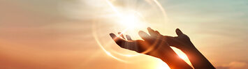 A person's hands reach, palm upward, toward the sky. A ball of light cast from the setting sun sits just above them.