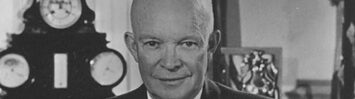 President Eisenhower behind his desk in the Oval Office, holding some papers, with his eyeglasses resting in front of him.
